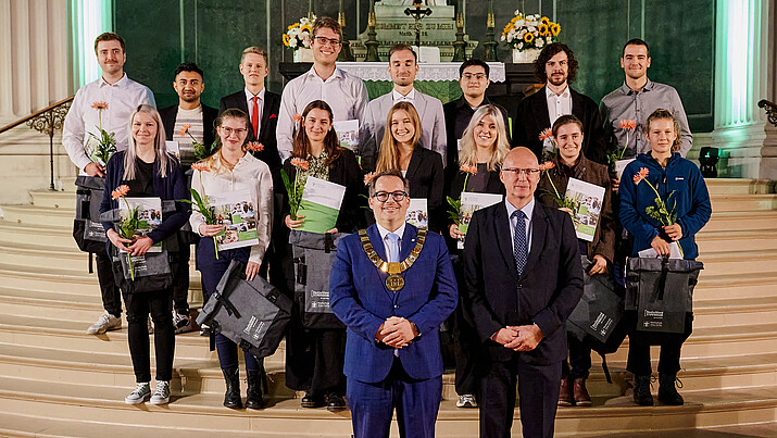 Eine Gruppe von Deutschlandstipendiatinnen und Deutschlandstipendiaten steht auf den Stufen der Kanzel der Johanniskirche. Im Vordergrund stehen der Rektor und Hochschulratsvorsitzende nebeneinander.