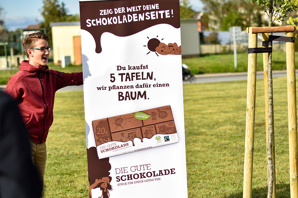 Männliche Person steht auf einer grünen Wiese neben einem Baum und neben einem Roll Up mit Werbebotschaft drauf.