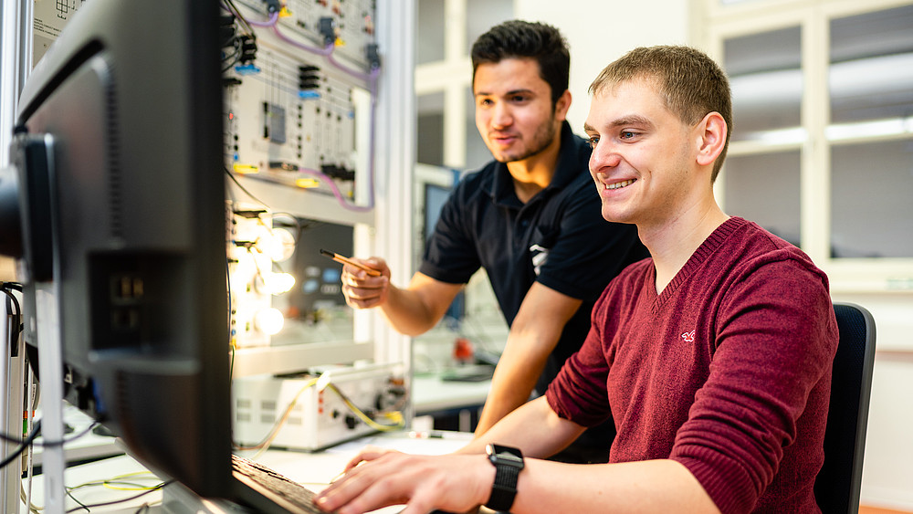 Zwei Studenten im Elektrotechniklabor, einer sitzt am Rechner, der andere steht dahinter und deutet auf den Bildschirm.