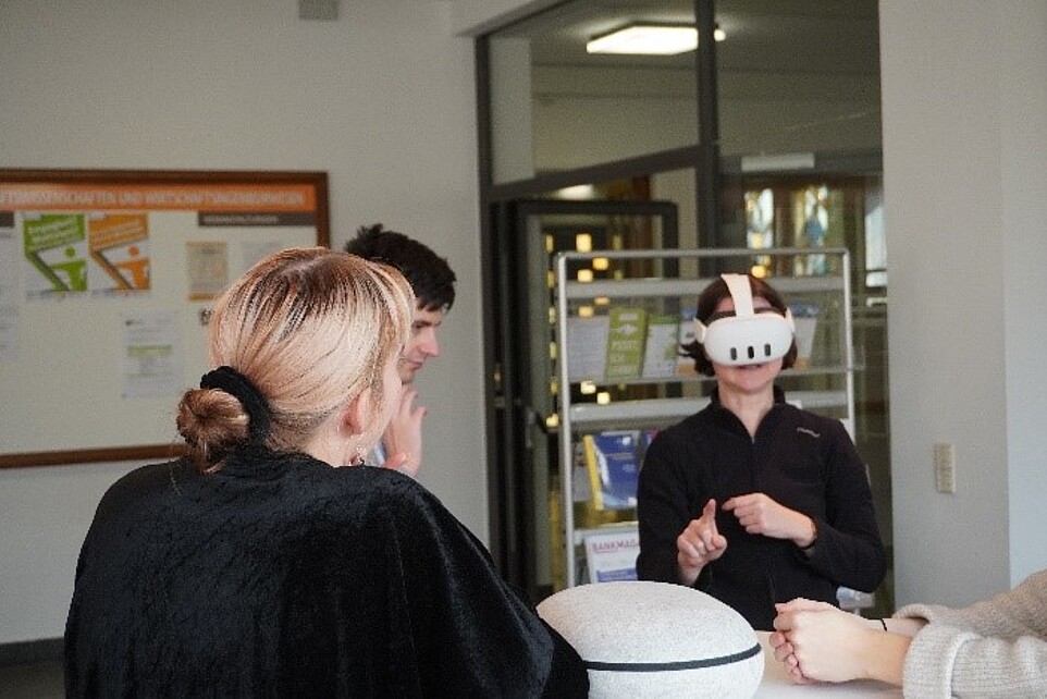 Am Mixed Reality Technologiestand probiert eine Mitarbeiterin eine weiße Mixed Reality-Brille auf und wird dabei von anderen Personen angesehen.