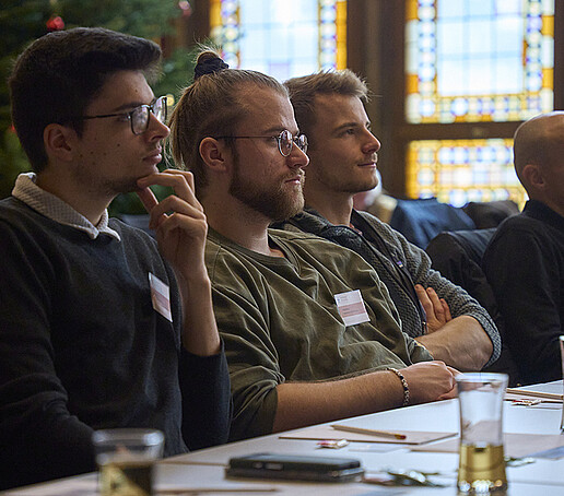 Teilnehmende sitzen im Publikum und richten ihren Blick nach vorn zu den Vortragenden.