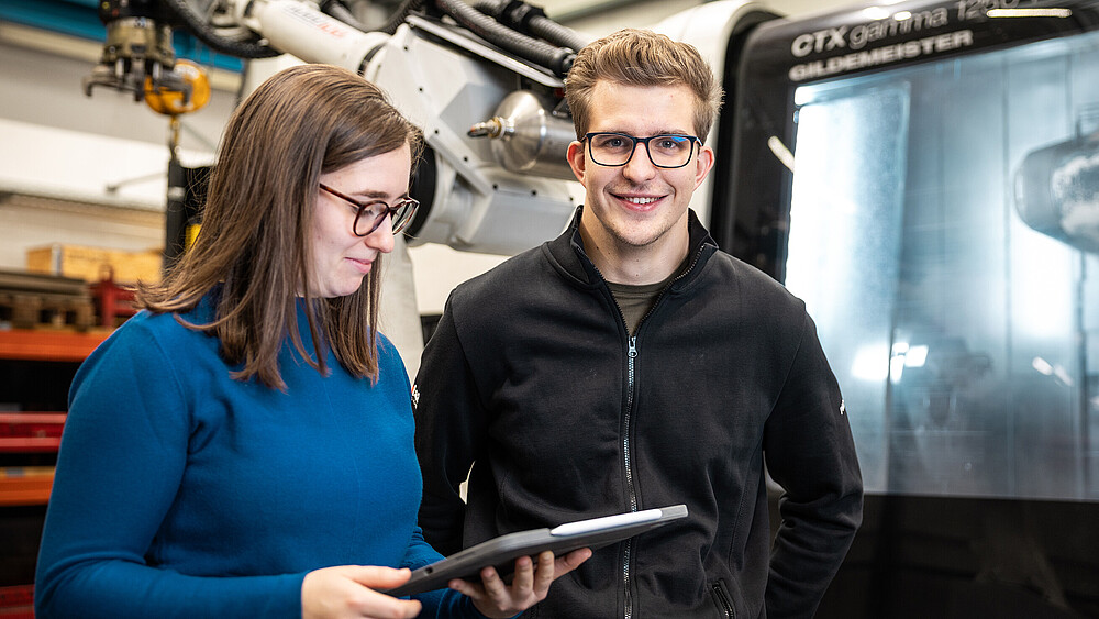 Zwei Studierende stehen an einer Anlage mit mechanischem Greifarm. Die Studentin schaut auf ein Tablet.