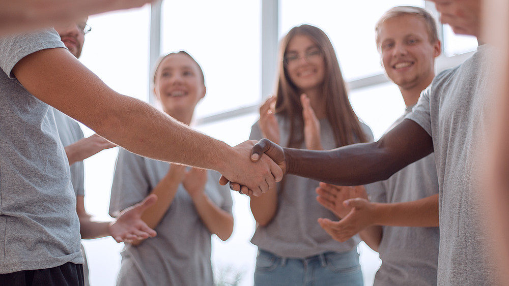 Zwei Gruppenmitglieder schütteln Hande, junge Studierende stehen dabei und applaudieren.