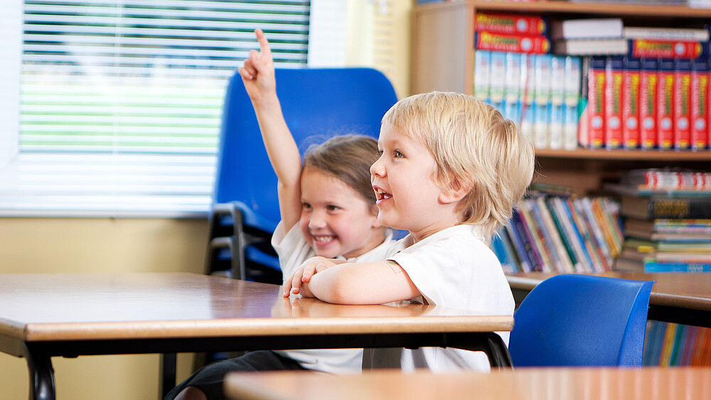 Zwei Grundschulkinder sitzen am Schultisch. Das Mädchen meldet sich.