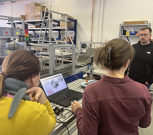 Ein Mann erklärt 2 anderen Personen die Cobot Versuchslinie