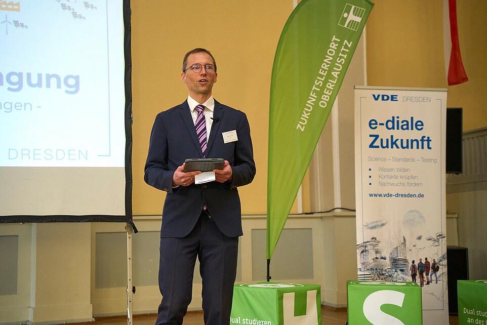 Redner mit Tablet auf Bühne vor Projektion und Veranstaltungsfahnen.