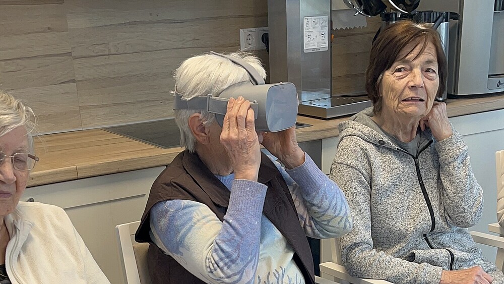 Eine alte Dame trägt eine VR-Brille, zwei alte Damen sitzen links und rechts daneben.