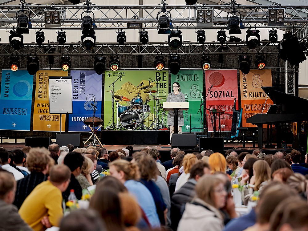 Blick auf eine Bühne mit einer Sprecherin, davor viel Publikum.