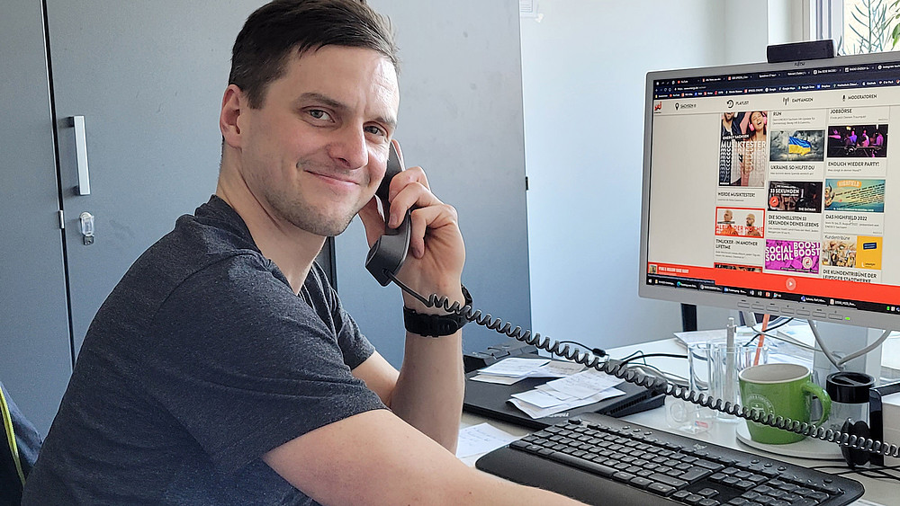Rico Winkler lächelt in die Kamera, er sitzt am Schreibtisch, hält einen Telefonhörer in der Hand und auf seinem PC-Bildschirm im Hintergrund ist die Webseite von Radio Energy Sachsen eingeblendet.