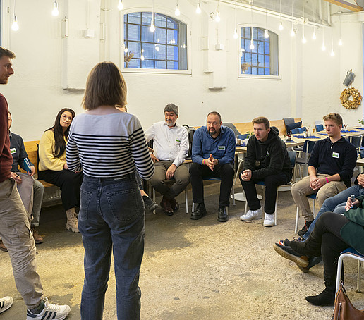 Im Saal sitzen Teilnehmende auf Stuhlreihen und hören zwei stehenden Teilnehmenden zu.