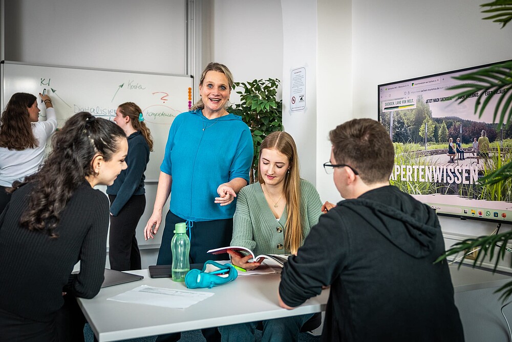 Das Bild zeigt eine lebendige Unterrichtsszene in einem modernen Klassen- oder Seminarraum. Im Vordergrund sitzen drei junge Erwachsene (zwei Frauen, ein Mann) an einem Tisch und arbeiten gemeinsam mit einem aufgeschlagenen Buch. Eine Frau mittleren Alters, vermutlich die Lehrkraft, steht lächelnd daneben und erklärt oder kommentiert etwas. Die Atmosphäre wirkt freundlich und engagiert.  Im Hintergrund arbeiten zwei weitere junge Frauen an einem Whiteboard, auf dem Begriffe wie „KI“, „Kosten“, „Digitalisierung“ und „Kompetenz“ notiert sind – ein Hinweis auf ein Thema rund um Digitalisierung oder Tourismusbildung.  Rechts im Bild befindet sich ein Bildschirm, auf dem eine Website mit dem Titel „EXPERTENWISSEN“ zu sehen ist. Oben erkennt man Begriffe wie „Sachsen. Land von Welt“ und einen Aufruf zur Reise nach Sachsen – es scheint sich um eine Präsentation oder Schulung zum Thema Tourismus in Sachsen zu handeln.  Zusätzliche Details:  Auf dem Tisch liegen ein Laptop, eine Wasserflasche, ein Mäppchen und Unterlagen.  Die Umgebung ist hell, freundlich und mit Pflanzen dekoriert.  An der Wand hängt ein Schild mit Verhaltensregeln (z. B. „Kein Essen & Trinken“).