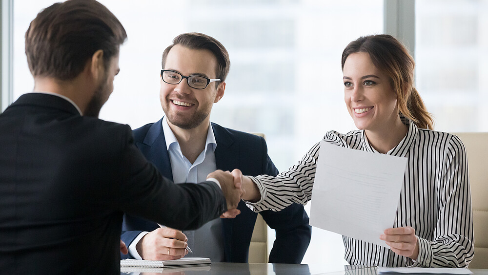 An einem Tisch reicht ein Mann im Anzug einer Frau die Hand, die gegenüber sitzt und einen Zettel in der anderen Hand hält.