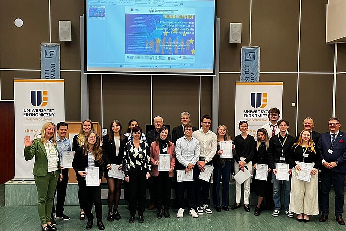 Eine große Gruppe Konferenzteilnehmender steht nebeneinander zum Gruppenfoto vor einer Beamerleinwand.