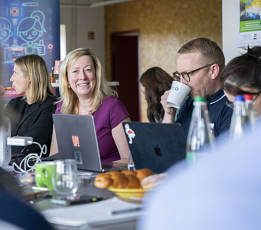 Die Prorektorin sitzt am Tisch und lächelt, ihr Tischnachbar trinkt einen Schluck aus einer Tasse.