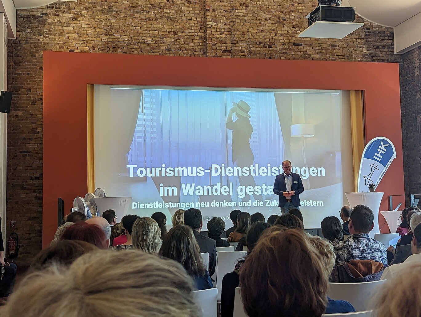 Blick aus dem Publikum auf eine Bühne, auf einen Mann mit Mikrofon 