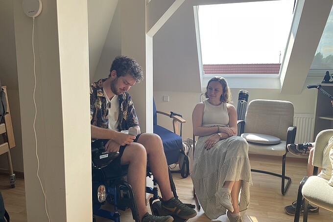 Ein junger Mann testet einen Rollstuhl und eine junge Frau sieht ihm dabei zu