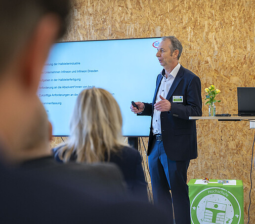 Ein Vortragender steht zwischen Bildschirm und Stehtisch und ist dem Publikum zugewandt.