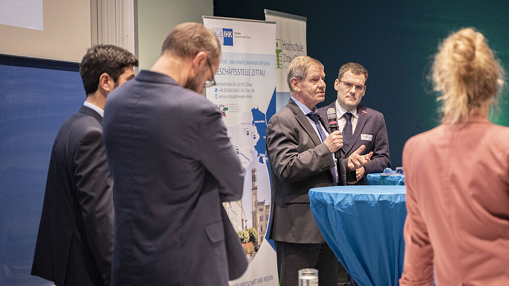 Eine Gruppe von Veranstaltungsteilnehmenden steht im Veranstaltungssaal zusammen und hören einem Vortragenden zu.