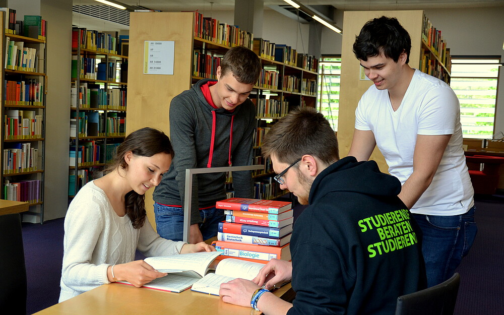 Vier Studierende befinden sich in der Bibliothek. Ein SbS-Student zeigt auf einen Text, der vor ihm auf dem Tisch liegt. Die anderen hören interessiert zu. Eine Studentin blättert in einem vor ihr aufgeschlagenen Heft. 
