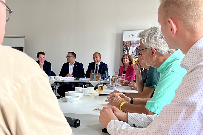 Die Gesellschafterinnen und Gesellschafter sitzen in einem Seminarraum an Tischen und besprechen sich.