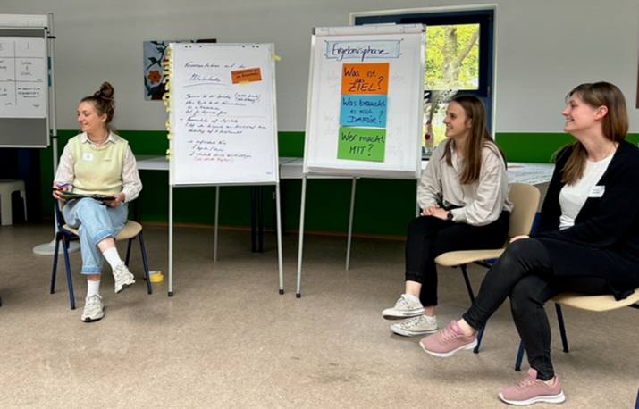 Drei Studentinnen sitzen im Halbkreis vor Aufstellern, die beschriftet und teilweise mit Zetteln beklebt sind.