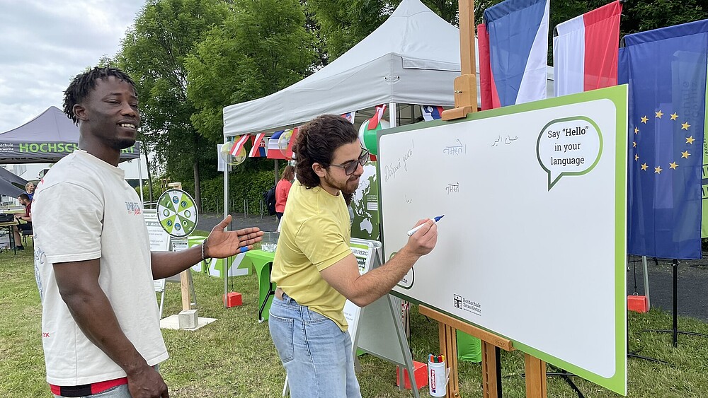 Zwei internationale Studierende stehen vor einer Tafel. Einer von ihnen schreibt mit einem Stift einen Gruß auf die Tafel.