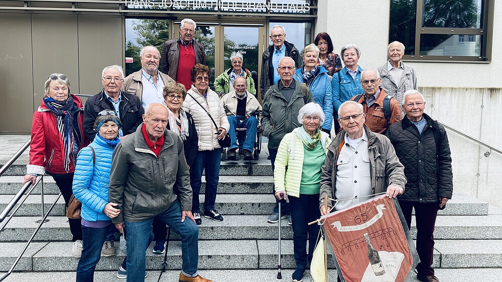 Eine Gruppe ehemaliger Studierender steht auf den Treppenabsätzen vor Haus eins. Ein älterer Herr hält eine Studiumsflagge in der Hand. Alle schauen in die Kamera.