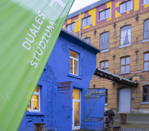 Ein Backsteingebäude steht im Hintergrund und links davor ein blaues Haus mit weißen Fensterläden. Im Vordergrund weht eine Beachflag mit der Aufschrift "Duales Studium".
