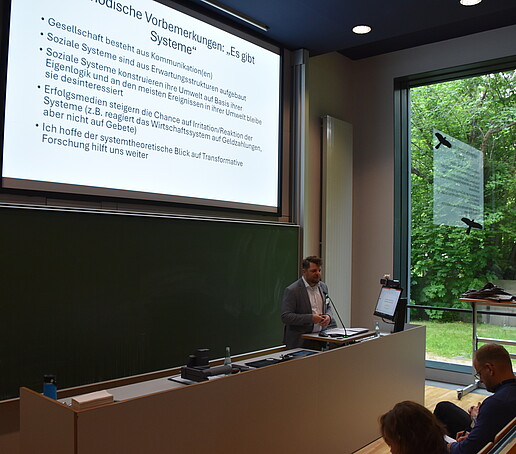 1 Redner steht am Pult im Hörsaal vor einer Präsentation