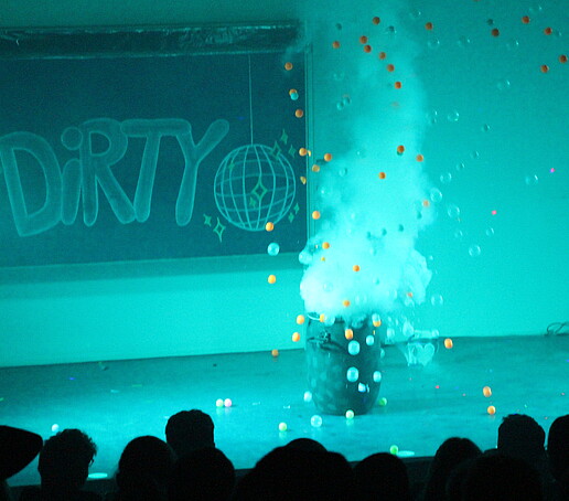 Im türkisen Licht fliegen bunte Bälle aus einer Tonne auf der Bühne.