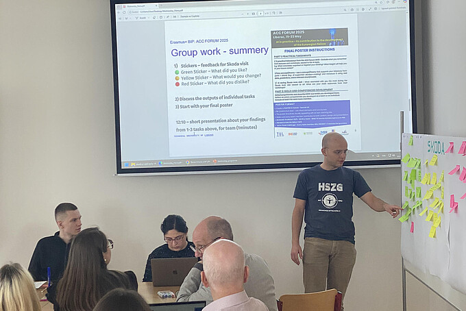 Ein Student erklärt einer Gruppe etwas an einem Aufsteller mit bunten Karten. Im Hintergrund erscheint eine Präsentation auf einem Bildschirm.