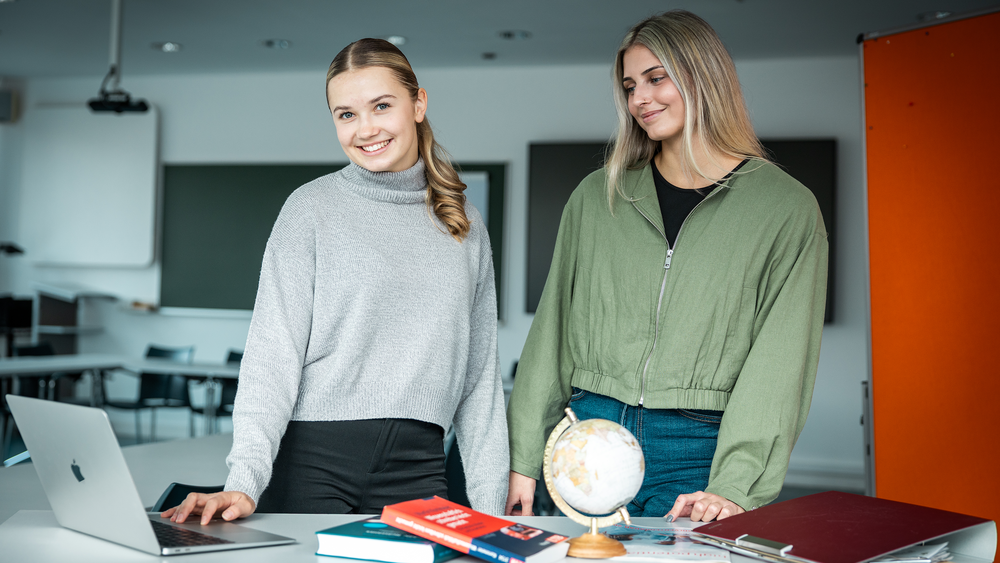 Zwei Studentinnen stehen an einem Tisch mit aufgeklappten Laptop. Vor ihnen liegen Studienmaterialien.