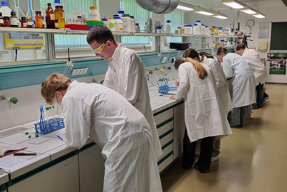 Schüler und Schülerinnen in weißen Kitteln stehen im Labor und experimentieren.