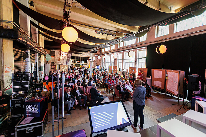 Publikum sitzt in einem großen Raum und hört einem Vortrag zu, im Hintergrund eine große Leinwand.