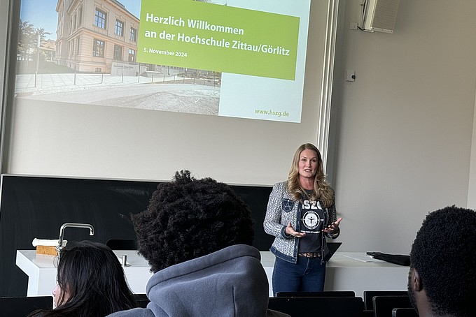 Inna Hauf steht im vorderen Bereich eines Hörsaals und spricht zu einer Gruppe internationaler Studieninteressierter des Rahn Studienkollegs Leipzig. Im Hintergrund ist eine Leinwand zu sehen, auf der eine Präsentation mit dem Titel „Herzlich Willkommen an der Hochschule Zittau/Görlitz“ angezeigt wird. Die Zuhörer sitzen in Reihen vor ihr und folgen aufmerksam ihrer Begrüßung.