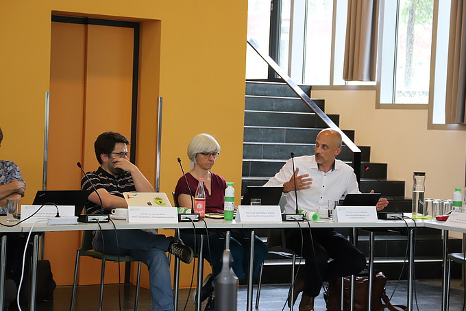 Zwei Dekane und eine Dekanin sitzen an einem langen Tisch in der Aula und beraten sich.