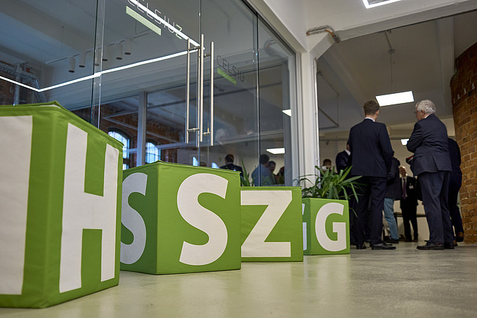 Grüne, würfelförmige Sitzkissen mit großen Buchstaben „H“, „S“, „Z“, „G“ in einem modernen Raum, im Hintergrund stehen Personen.