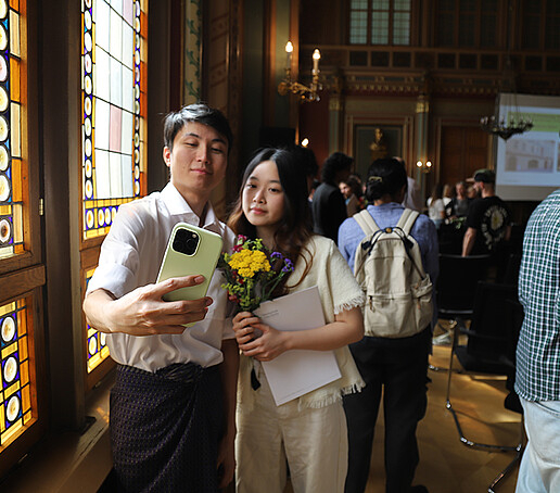 Eine Absolventin und ihre Begleitung machen neben den bunten Glasfenstern ein Selfie.