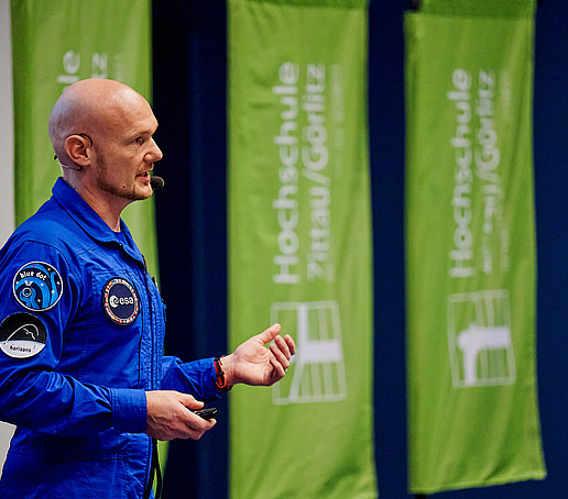 Alexander Gerst steht auf dem Podium im Hörsaal, im Hintergrund stehen drei Hochschulfahnen.