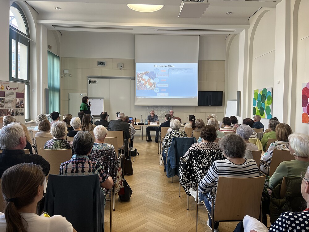 Viele Menschen sitzen in einem Saal und schauen auf eine Leinwand