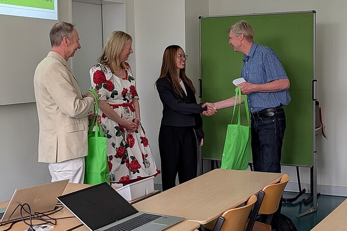 Steffen Zücker bedankt sich im Namen der Fakultät Wirtschaftswissenschaften und Wirtschaftsingenieurwesen und Hochschule Zittau/Görlitz bei den Referierenden und überreicht Frau Nguyen einen grünen HSZG-Beutel. Daneben stehen die Prorektorin und Dr. Schneider im Seminarraum vor der ersten Sitzreihe.