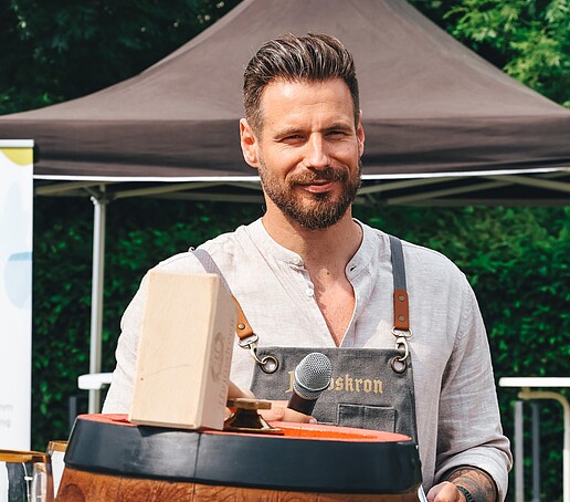 Philipp Knebel steht hinter einem Bierfass und vor einem Pavillon und lächelt.