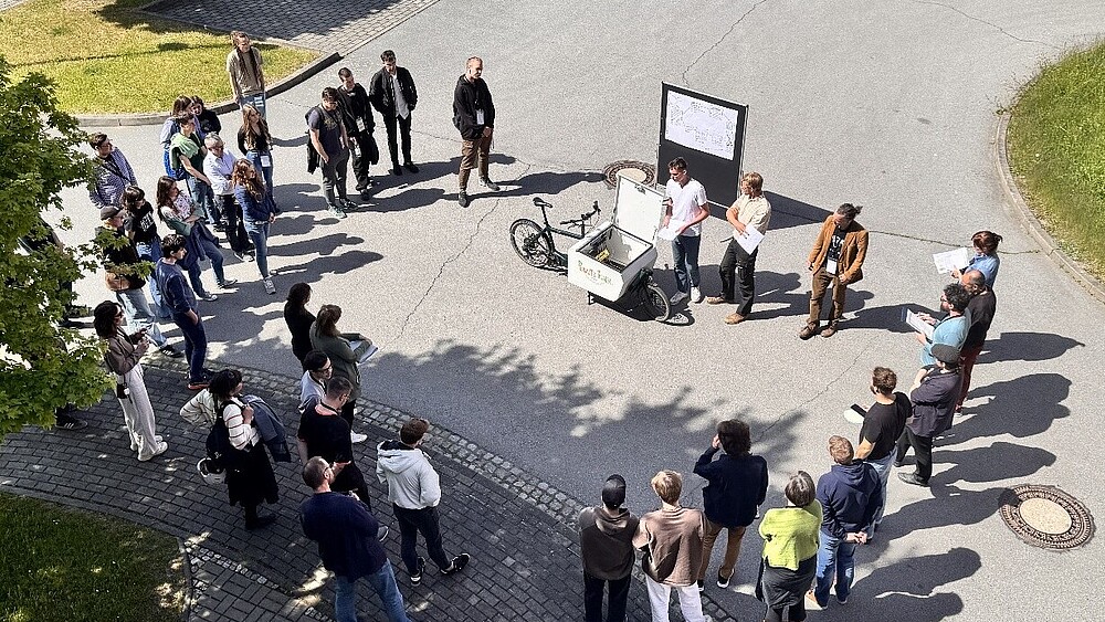 Eine Gruppe Menschen stehen auf einem Hof im Kreis. An der oberen Seite steht ein Fahrrad und ein Blackboard.