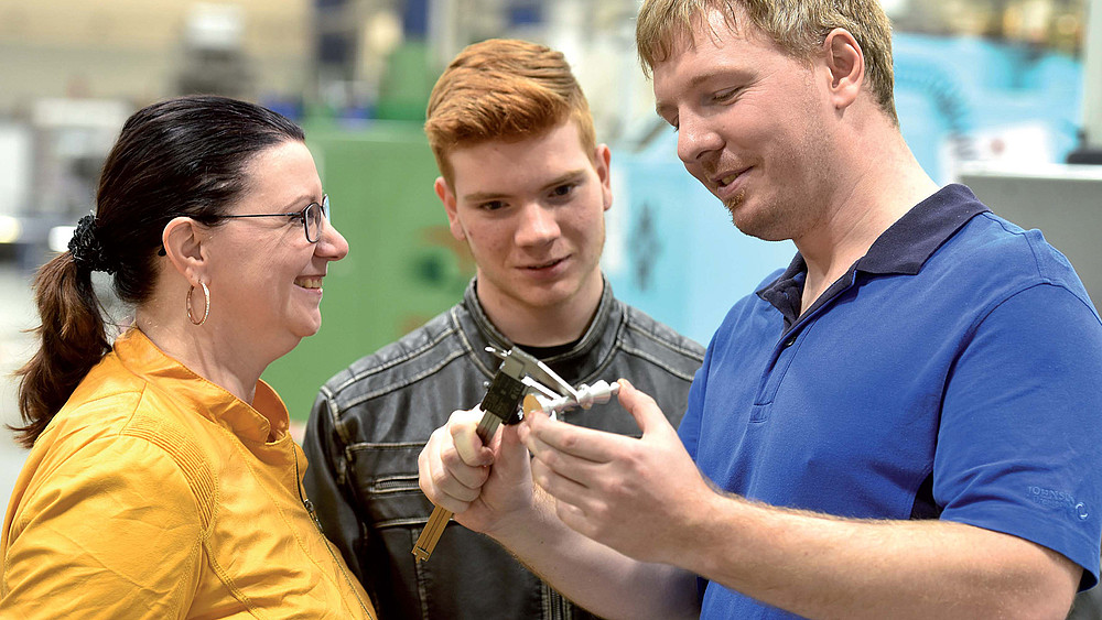 Ein Mann im blauen Poloshirt vermisst einen Gegenstand ein jüngerer Mann und eine Frau schauen dabei zu.