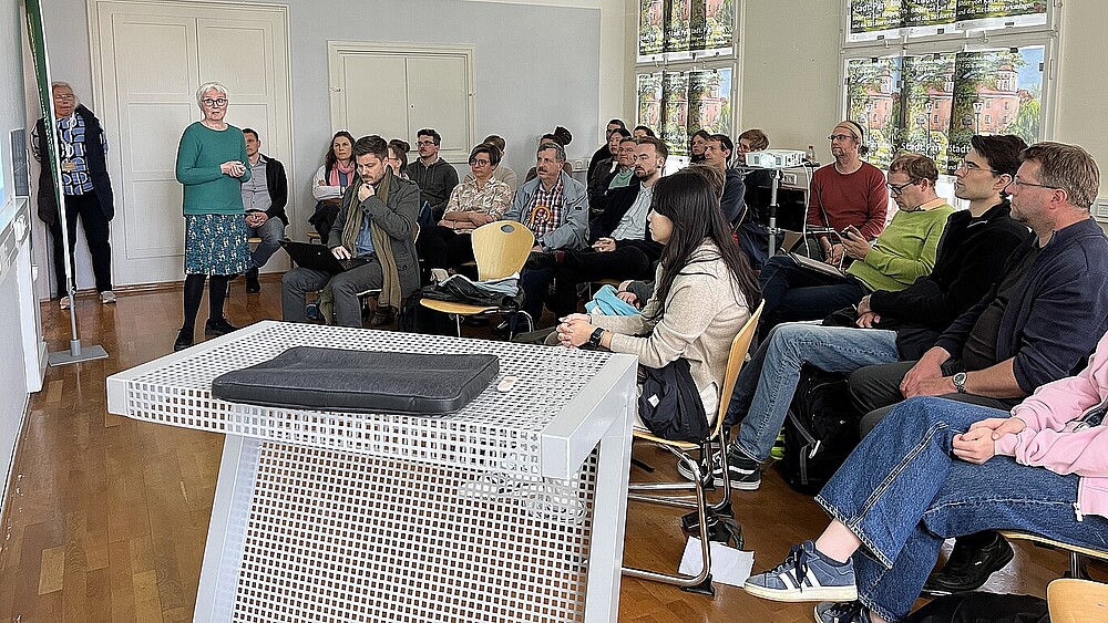 Teilnehmende eines Fachaustauschs sitzen auf Stuhlreihen und hören einer Dozentin zu.