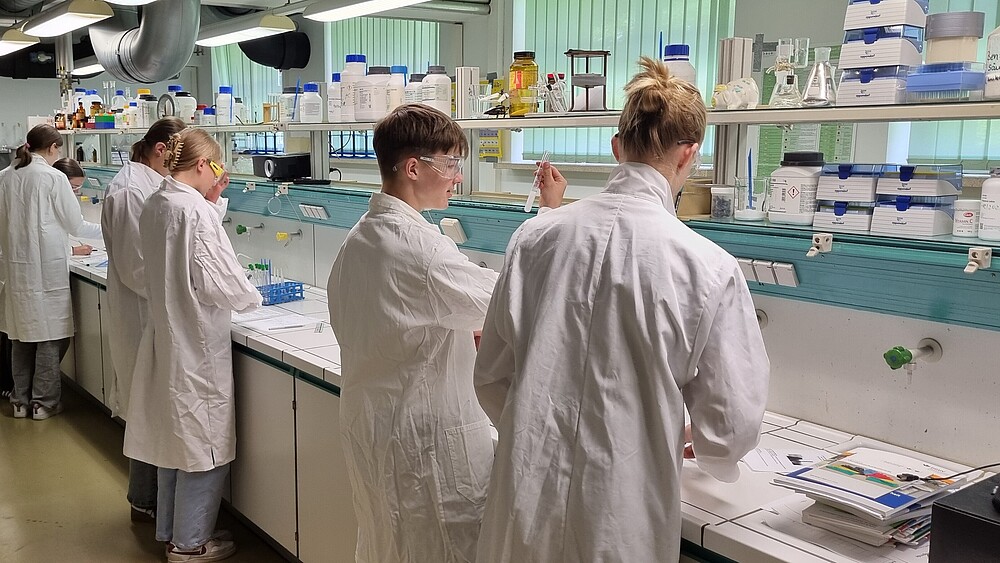 Schüler und Schülerinnen in weißen Kitteln stehen im Labor und experimentieren.