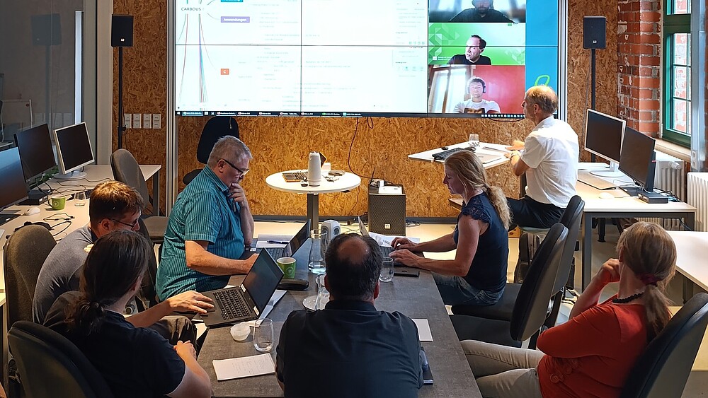 Menschen sitzen an Tischen im Celsiuz, im Hintergrund steht eine große digitale Leinwand, auf der Präsentationen abgebildet und Personen live per Webcam zugeschaltet sind.