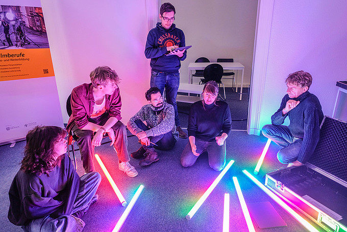 Kursteilnehmende und Dozentin sitzen auf dem Boden mit Lichtröhren. Die Dozentin demonstriert die Lichteinstellung mittels Tablet.