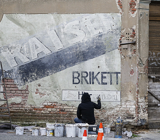 Ein Filmset bestehend aus einer alten Steinmauer. Davor kniet jemand und bemalt die Mauer.