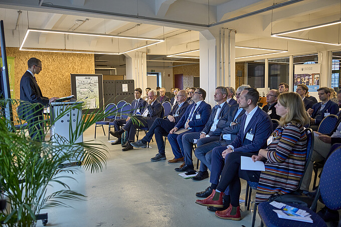 Publikum hört einem Vortragenden zu, der vor einem Rednerpult steht; ein großes Plakat oder Diagramm steht im Hintergrund.